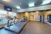 This single-level fitness center is thoughtfully designed to accommodate a comprehensive workout experience. The space is characterized by a distinctive tray ceiling with a dark-painted recessed field accented by crown molding, creating a polished, structured aesthetic throughout. Recessed and flush-mounted lighting fixtures provide consistent illumination across the entire floor plan.

The cardio zone is generously outfitted with multiple treadmills, elliptical trainers, and stationary and spin bikes, several of which are positioned along a windowed wall that offers views of an outdoor fenced area, allowing natural light to fill the space. A wall-mounted flat-screen television is positioned for easy viewing during workouts.

The strength training area includes a multi-station cable machine, a full rack of dumbbells in varying weights, medicine balls, resistance bands, and a weight bench. Large mirrors along one wall visually expand the space and assist with form correction.

The flooring throughout is durable rubber, ideal for heavy equipment and high-impact use. The neutral beige walls and clean white trim contribute to a bright, functional environment. Posted fitness rules and emergency signage reflect a well-maintained, safety-conscious facility.