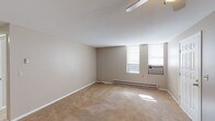 This single-floor unit offers a straightforward, functional layout with distinct living, sleeping, and kitchen areas. The main living space is generously proportioned, finished with neutral beige carpeting and warm taupe walls that create a calm, understated atmosphere. A ceiling fan with integrated lighting provides overhead illumination, while multiple windows along the exterior wall allow natural light to filter in throughout the day. Baseboard heating runs along the perimeter walls, and a wall-mounted air conditioning unit ensures year-round climate control.

The layout, as visible in the 3D floor plan, separates the living area from the kitchen and additional rooms via interior doorways. A closet with bifold doors is conveniently situated near the main living space, offering practical storage. The secondary room features darker carpeting and a similar neutral wall palette with a single window.

The kitchen is equipped with oak cabinetry above and below white laminate countertops, a stainless steel sink, a white electric range, and a dishwasher. Dark hardwood-style flooring grounds the kitchen area, providing a contrast to the carpeted rooms. The overall design is clean and utilitarian, suited for comfortable everyday living.