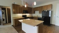 This single-floor space showcases a thoughtfully designed kitchen that serves as the heart of the home. The kitchen is anchored by a spacious central island topped with elegant granite countertops in warm golden and cream tones, providing ample prep space and a natural gathering point. Matching granite surfaces extend throughout the kitchen, complementing the rich, medium-toned wood cabinetry that lines the walls and reaches toward the dramatically vaulted ceilings above. The full suite of black appliances — including a French door refrigerator with an ice and water dispenser, an electric range, over-the-range microwave, and dishwasher — lends a sleek, cohesive look to the space. Dark hardwood flooring grounds the kitchen and flows throughout, adding warmth and contrast to the lighter countertops and cream-colored walls. Pendant lighting hangs gracefully above the island, providing focused illumination while enhancing the overall ambiance. The soaring vaulted ceilings amplify the sense of openness and volume, making the kitchen feel expansive and airy. Under-cabinet lighting adds a refined finishing touch, highlighting the countertops and creating a welcoming, well-lit environment suited for both everyday cooking and entertaining.