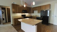 This single-floor space showcases a thoughtfully designed kitchen that serves as the heart of the home. The kitchen is anchored by a spacious central island topped with elegant granite countertops in warm golden and cream tones, providing ample prep space and a natural gathering point. Matching granite surfaces extend throughout the kitchen, complementing the rich, medium-toned wood cabinetry that lines the walls and reaches toward the dramatically vaulted ceilings above. The full suite of black appliances — including a French door refrigerator with an ice and water dispenser, an electric range, over-the-range microwave, and dishwasher — lends a sleek, cohesive look to the space. Dark hardwood flooring grounds the kitchen and flows throughout, adding warmth and contrast to the lighter countertops and cream-colored walls. Pendant lighting hangs gracefully above the island, providing focused illumination while enhancing the overall ambiance. The soaring vaulted ceilings amplify the sense of openness and volume, making the kitchen feel expansive and airy. Under-cabinet lighting adds a refined finishing touch, highlighting the countertops and creating a welcoming, well-lit environment suited for both everyday cooking and entertaining.