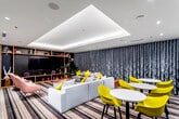 This single-level cinema and lounge space is designed with a contemporary aesthetic that balances functionality and visual flair. The room is anchored by a large projection screen and a dedicated cinema wall featuring the word "CINEMA" in bold neon lettering, flanked by oversized artwork and a built-in wooden shelving unit displaying books, decorative objects, and a flat-screen television. Ambient lighting is a defining architectural feature, with a recessed tray ceiling fitted with continuous LED cove lighting that casts a warm, even glow throughout the space. Recessed downlights complement this effect, creating a layered and inviting atmosphere. Floor-to-ceiling dark, textured drapes line an entire wall, adding depth and a dramatic cinematic quality to the room. The flooring features a bold striped carpet that grounds the space and ties together the varied seating zones. Furnishings are arranged to encourage both social interaction and screen viewing. A large modular grey sofa is accented with vibrant yellow and black-and-white patterned cushions, while a pair of plush pink accent chairs provide a softer counterpoint. Multiple round white café tables paired with chartreuse yellow chairs offer casual seating. A low round coffee table and a small concrete side table complete the layout, making this a versatile and well-appointed communal entertainment space.