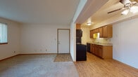 This single-floor unit offers a practical and comfortable layout designed for everyday living. The open living area is finished with plush carpet and flows seamlessly into a secondary room fitted with a ceiling fan, creating a versatile space suitable for a variety of uses. Light wood-look flooring defines the transition into the kitchen and entryway areas, providing a clean visual contrast.

The kitchen is efficiently designed with warm-toned wood cabinetry both above and below the counters, a light laminate countertop, a double-basin sink, and black appliances including a refrigerator, range, and dishwasher. Overhead lighting keeps the kitchen well-lit throughout the day.

Two bedrooms offer comfortable proportions, each featuring neutral carpet, white walls, warm wood trim, and windows dressed with horizontal blinds that allow for natural light and privacy control. The rooms provide a calm, understated atmosphere well-suited for rest or a home office setup.

The bathroom includes a full-sized soaking tub with a built-in surround featuring integrated shelving, finished in clean white. Throughout the unit, consistent use of wood-toned trim, neutral tones, and practical finishes contribute to a cohesive and functional living environment.