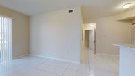 This single-storey space presents a clean, minimalist interior defined by light-toned walls and large white ceramic floor tiles that extend throughout, creating a cohesive and airy feel. The main open area is generously sized, with smooth beige walls, white baseboards, and a ceiling-mounted HVAC vent indicating a central air system. The space transitions into a corridor that leads to at least one additional room, visible through a white panel door at the end of the hallway. Overhead lighting in the hallway provides practical illumination, while the overall color palette — neutral creams and whites — contributes to a bright, clean atmosphere. The layout appears straightforward and functional, with electrical outlets positioned conveniently along the walls. The absence of clutter and the uniform flooring throughout reinforce a sense of openness and ease of movement between areas. The space is well-suited for a variety of uses, offering a blank canvas that can be adapted to different configurations and purposes.