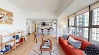 This single-floor residence showcases a thoughtfully designed open-concept layout that seamlessly connects the living, dining, and kitchen areas. Light wide-plank hardwood flooring flows throughout, complementing the crisp white walls and lending a warm, contemporary feel to the entire space.

The living area is anchored by expansive floor-to-ceiling windows that frame impressive urban skyline views, flooding the room with natural light. The kitchen is a standout feature, equipped with white flat-front cabinetry accented by matte black hardware, a gas range, stainless steel refrigerator, subway tile backsplash, and open wood shelving. A generous island with a white quartz countertop provides seating for two and serves as the social hub of the space.

Both bedrooms are generously proportioned, each featuring large windows with city views and the same warm hardwood flooring. The two bathrooms share a cohesive design language, featuring bold black hexagonal floor tiles paired with white honeycomb wall tiles. One bathroom includes a glass-enclosed walk-in shower, while the other offers a full soaking tub with a shower combination, both finished with modern floating vanities and wood accents.