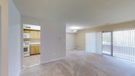 Unit 108 is a single-floor residence designed with a practical and open layout. The spacious living area is finished with neutral carpet flooring and light-toned walls, creating a calm and inviting atmosphere. Large vertical blinds frame a sliding glass door that brings in natural light and provides access to the outdoors, enhancing the sense of openness in the main living space. A ceiling-mounted light fixture adds to the room's even illumination. Adjacent to the living area, the kitchen is separated by a half-wall pass-through that maintains a connected feel between the two spaces. The kitchen features light wood cabinetry, offering ample storage, and is complemented by tile flooring that contrasts with the carpeted living areas. White appliances contribute to the clean, straightforward aesthetic of the kitchen. Throughout the unit, white baseboards and neutral wall tones provide a cohesive and understated design. The single-floor plan ensures ease of movement throughout the space, making it well-suited for everyday living. Overall, Unit 108 offers a functional, well-lit, and comfortable environment with a simple, classic interior style.