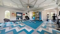 Located in Saint Petersburg, Florida, this fitness center offers a spacious and well-appointed exercise environment designed to support a wide range of workout routines. The room makes an immediate architectural impression with its ornate coffered ceilings, detailed molding, and a central ceiling fan — elements that lend the space a refined, upscale character uncommon in typical gym settings. Abundant natural light pours in through multiple arched and French-style windows and glass doors, creating a bright and inviting atmosphere throughout.

The flooring features a bold teal, white, and gray chevron-patterned carpet tile design that adds visual energy to the space. Cardio equipment is well-represented, with a dedicated row of treadmills positioned along a wall-mounted flat-screen TV with a teal accent backsplash, as well as StairMaster machines and ellipticals. A functional training station holds an array of resistance bands, suspension trainers, and colorful stability balls, supporting dynamic and flexibility-focused workouts. Adjustable benches, rowing machines, and a wall-mounted TV with a built-in media niche round out the amenities. French doors along multiple walls provide views of the surrounding outdoor greenery and easy access to the exterior, enhancing the open, airy feel of this thoughtfully designed fitness facility.