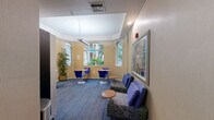 Located at 6001 SW 70th St in South Miami, FL, this single-room common area serves as a versatile shared space combining business center functionality with comfortable lounge amenities. The room features warm beige walls, a tray ceiling with recessed lighting, and a patterned blue carpet that anchors the space with a cohesive, professional aesthetic.

Arched windows along multiple walls flood the interior with natural light and offer views of the surrounding tropical landscaping, including lush palm trees and greenery. A tall potted bamboo plant adds a natural, decorative touch to the interior.

The space is thoughtfully organized into distinct functional zones. A built-in white desk runs along one wall, providing two workstations complete with ergonomic office chairs — ideal for productivity tasks. A snack and beverage vending machine is conveniently positioned nearby.

For more casual use, round wooden café tables paired with vibrant blue chairs offer informal seating arrangements, while a lounge area features upholstered blue chairs and a cushioned bench centered around a small side table. Framed artwork, including a colorful abstract print, decorates the walls and adds visual interest. The overall ambiance is bright, welcoming, and practical, making this space well-suited for both focused work and relaxed socializing.