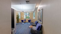 Located at 6001 SW 70th St in South Miami, FL, this single-room common area serves as a versatile shared space combining business center functionality with comfortable lounge amenities. The room features warm beige walls, a tray ceiling with recessed lighting, and a patterned blue carpet that anchors the space with a cohesive, professional aesthetic.

Arched windows along multiple walls flood the interior with natural light and offer views of the surrounding tropical landscaping, including lush palm trees and greenery. A tall potted bamboo plant adds a natural, decorative touch to the interior.

The space is thoughtfully organized into distinct functional zones. A built-in white desk runs along one wall, providing two workstations complete with ergonomic office chairs — ideal for productivity tasks. A snack and beverage vending machine is conveniently positioned nearby.

For more casual use, round wooden café tables paired with vibrant blue chairs offer informal seating arrangements, while a lounge area features upholstered blue chairs and a cushioned bench centered around a small side table. Framed artwork, including a colorful abstract print, decorates the walls and adds visual interest. The overall ambiance is bright, welcoming, and practical, making this space well-suited for both focused work and relaxed socializing.
