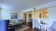 This single-story residence showcases an open-concept design that connects the living, dining, and kitchen areas into one cohesive, flowing space. Rich, dark hardwood flooring runs throughout, providing a warm contrast to the crisp white walls and light-toned ceilings, which feature a subtle texture overhead. The living area is generously proportioned, offering ample room for comfortable seating arrangements, and is anchored by a patterned area rug. A large framed artwork adds a vibrant focal point to the main wall, enhancing the space's aesthetic appeal. A structural column elegantly divides the living zone from the dining area, where bar-height seating lines a half-wall partition. This partition opens directly to the kitchen, which features warm wood cabinetry and a bright, functional layout. Natural light filters through the space, contributing to its airy ambiance. A hallway visible from the main area leads to additional rooms, including a bathroom with a curtained shower, suggesting a well-organized and practical floor plan throughout. The overall aesthetic is modern, clean, and welcoming.