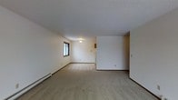 This single-floor residence offers a straightforward and functional layout designed for comfortable everyday living. The main living area is generously proportioned, featuring wall-to-wall beige carpeting that adds warmth underfoot and contributes to the overall neutral, understated aesthetic. White walls throughout the space provide a clean, bright backdrop that can easily accommodate a variety of furnishing styles and décor preferences. The textured ceiling adds a subtle architectural detail consistent across the interior. A window in the living area allows natural light to filter into the space, helping to brighten the room. A ceiling-mounted light fixture provides additional illumination. Baseboard heating units run along the lower perimeter of the walls, offering a practical and efficient heating solution. The open floor plan creates a sense of continuity and flow between areas, with a partial wall defining a transitional zone that leads toward what appears to be an adjoining room or kitchen area. Warm wood-toned baseboards frame the carpeted floors, providing a gentle contrast to the white walls. Overall, the space presents a clean, move-in ready environment with a calm and neutral ambiance well-suited for a variety of living arrangements.