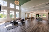This single-floor community clubhouse is defined by its dramatic architectural character and thoughtfully designed gathering spaces. The main lounge area commands attention with soaring double-height ceilings painted in a striking teal, from which a statement globe chandelier is suspended. A full-height stacked stone accent wall anchors the room, featuring a linear electric fireplace and a mounted flat-screen TV, creating a warm and inviting focal point. Wide-plank wood-look flooring flows throughout the entire space, unifying the various zones seamlessly. Rows of large windows flood the interior with abundant natural light while framing verdant outdoor views, enhancing the open and airy ambiance. The fully equipped kitchen is a standout feature, showcasing bold forest-green cabinetry paired with white upper cabinets, a textured tile backsplash, stainless steel appliances, and a generous white quartz island with bar seating beneath a trio of elegant glass pendant lights. Adjacent to the kitchen, the dining area accommodates multiple long communal tables and is appointed with soft sage-green walls, linen curtains, and decorative wall art. Black-framed French doors open directly to an outdoor patio, seamlessly connecting the interior to the surrounding landscape.
