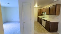 This single-floor space presents a functional and straightforward layout connecting the kitchen and living areas. The kitchen is equipped with dark wood cabinetry spanning both upper and lower sections, providing ample storage. Light-colored countertops offer a contrasting workspace, and a built-in sink with a dishwasher adds practical convenience. The kitchen flooring consists of a neutral, tile-style surface that is easy to maintain. Overhead, a flush-mount ceiling light provides direct illumination, and a textured ceiling is visible throughout. Adjacent to the kitchen, a doorway leads to a carpeted living area, which features neutral-toned carpet that contributes a softer, more comfortable feel underfoot. A tall, white interior door is prominently positioned at the transition point between the two areas, likely providing access to a closet or utility space. The walls throughout are painted in a clean, neutral white, keeping the overall aesthetic simple and versatile. The space is compact yet efficiently organized, with a clear separation between the kitchen's hard flooring and the living area's carpeted surface, defining each functional zone distinctly.