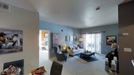 This single-floor residence offers a thoughtfully designed living space that balances comfort and contemporary style. The living room serves as the heart of the home, featuring plush wall-to-wall carpeting and cool blue-toned walls that create a calm, welcoming atmosphere. Large sliding glass doors dressed with elegant patterned drapes flood the space with natural light and provide direct access to an outdoor patio or balcony area, offering pleasant views beyond. The room is generously proportioned, comfortably accommodating a full seating arrangement including a sofa, wingback chairs, and an upholstered ottoman, all anchored by a decorative area rug. Curated wall art adds personality and visual interest throughout the space. An adjacent bedroom is visible through an open doorway, hinting at a well-connected and functional floor plan. The smooth white ceilings complement the overall clean aesthetic of the interior. Together, the design elements create a cohesive living environment that feels both stylish and livable, making this a practical yet attractive space for everyday living.