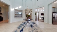 Villages 3Eighty is a thoughtfully designed single-floor clubhouse space that combines architectural elegance with modern amenities. The grand entry foyer sets an impressive tone immediately upon arrival, featuring soaring ceilings with arched detailing, large-format light tile flooring, and a bold blue-and-white abstract area rug anchored by a sleek glass-top accent table adorned with a dramatic floral arrangement. The prominently displayed Villages 3Eighty branding adds a polished, professional identity to the welcome space.

Beyond the foyer, the great room commands attention with its stunning coffered ceiling, rich hardwood flooring, a classic fireplace, and generous seating arrangements accented with gold and teal throw pillows. Tall windows dressed in neutral drapes flood the room with natural light, while arched doorways lend a timeless architectural character throughout.

The gourmet kitchen is a standout feature, appointed with granite countertops, mosaic tile backsplash, sage-toned cabinetry, stainless steel appliances including a professional-grade range, and a tiled island with bar seating—ideal for entertaining.

A separate dining area offers a glass-top table surrounded by white upholstered chairs, bathed in natural light from floor-to-ceiling windows. The overall ambiance is refined yet welcoming, blending contemporary furnishings with classic architectural details seamlessly.