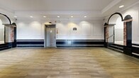 Located at 283 S Center St in Orange, NJ, this single-floor commercial space combines classic architectural character with a clean, updated interior. The open floor plan is finished with wood-look tile flooring throughout, creating a warm and cohesive aesthetic across the entire space. Walls are adorned with decorative rectangular panel moldings set against a crisp white and navy two-tone design, lending the interior a polished, formal ambiance. Crown molding runs along the ceiling perimeter, adding an elegant finishing touch. Recessed lighting provides even, bright illumination throughout. A standout feature is the ornate arched entryway, framed by a decorative wrought iron transom and accompanied by a set of steps with marble-accented columns, adding a sense of grandeur to the interior. A second arched window along one wall allows natural light to filter in, enhancing the brightness of the space. An elevator door is also present, indicating accessibility. The layout is open and flexible, making it well-suited for a variety of commercial uses such as retail, events, or professional services. Overall, the space presents a refined and inviting atmosphere rooted in distinctive architectural detail.