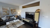 This single-floor space showcases a modern, sophisticated interior design that seamlessly blends functionality with refined aesthetics. The living area serves as the focal point of the space, anchored by a sleek, dark entertainment unit that houses a large flat-screen television and is complemented by accent cabinetry with warm yellow-gold tones, adding a striking contrast to the darker elements.

The living room features a plush charcoal gray sofa dressed with tasteful accent pillows, positioned around a contemporary gold-and-glass coffee table atop a bold chevron-patterned area rug in gray and white tones. Dark hardwood-style flooring runs throughout, grounding the space with warmth and depth.

Decorative details are carefully considered — a large abstract canvas with warm amber and gold tones adorns the wall, echoing the metallic accents found in the sculptural décor pieces and table lamp. Architectural interest is added by a recessed upper wall opening, lending a sense of dimension and openness to the living area.

The overall ambiance is chic and curated, with a cohesive neutral and gold color palette that creates an inviting yet polished atmosphere. The space reflects a high standard of interior design, making it both visually striking and comfortable for everyday living.