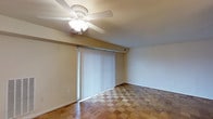 This single-floor space offers a straightforward and functional layout with several notable design details. The main living area showcases a classic parquet-style wood flooring laid in a checkered pattern, adding a distinctive visual texture to the room. Neutral, light-toned walls create a clean and airy atmosphere throughout the space.

A ceiling fan with an integrated light fixture is centrally positioned overhead, providing both air circulation and ambient lighting. The room is further brightened by a generously sized sliding glass door, dressed with full-length vertical blinds that allow for flexible light control and privacy. This door serves as a key architectural feature, drawing in natural light and creating a strong visual focal point along the wall.

Trim details including baseboards and door casings are finished in a crisp white, offering a subtle contrast against the warm wall tones. The overall ambiance of the space is modest and understated, presenting a neutral canvas well-suited for a variety of furnishing styles. The single-floor plan ensures easy accessibility and a cohesive flow throughout the living area. The combination of the parquet flooring, natural light access, and ceiling fan makes this a practical and comfortable environment.