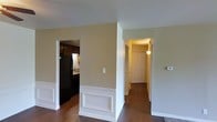 This single-story residence is organized around a central open area that connects multiple functional zones. The main living space features classic white wainscoting panels along the lower walls, adding a traditional architectural detail against the warm beige upper walls. Dark hardwood flooring runs consistently throughout, providing a rich contrast to the light-toned walls and white trim.

To one side, the kitchen is partially visible and equipped with dark cabinetry, a black refrigerator, and a sink positioned beneath under-cabinet lighting. The darker cabinet finish and appliances create a defined, utilitarian cooking space.

On the opposite side, a hallway leads to a white paneled entry door, warmly lit by a wall-mounted light fixture. The corridor is clean and straightforward, with the same hardwood flooring continuing through. The entryway provides a welcoming transition into the home.

Overall, the space maintains a cohesive aesthetic through consistent flooring, neutral wall colors, and white trim details. The layout is practical, with clear sightlines between the living area, kitchen, and entry, contributing to an efficient and connected single-floor living environment.