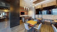 Located at 520 12th St S in Arlington, Virginia, this single-floor amenity space combines contemporary design with industrial-chic elements to create a dynamic and inviting environment. The space is anchored by a well-appointed kitchen bar featuring dark cabinetry, a built-in sink, under-counter refrigerators, a microwave, and a wall-mounted television set against a striking textured tile backsplash. Bold red accent walls with built-in display niches add vibrant contrast, while retro-inspired pendant lighting in red and white casts a warm, energetic glow over the natural wood communal dining table. Adjacent dining areas are furnished with wooden tables, upholstered chairs, and Edison bulb pendants, set against exposed brick walls that lend a warm, urban character to the space. A dedicated lounge area features a multi-screen media wall clad in reclaimed wood-style paneling, complemented by plush seating and bold artwork displayed against the exposed brick. Floor-to-ceiling windows and glass doors open onto a spacious outdoor terrace, where patio furniture and panoramic views of the Arlington skyline provide an exceptional setting for relaxation or entertaining. Throughout, the design balances industrial textures with modern finishes, creating a cohesive and sophisticated communal space.