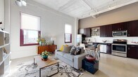 This urban loft showcases a thoughtfully designed open-concept layout that blends industrial character with modern comfort. Soaring ceilings with exposed ductwork and corrugated metal panels lend the space a distinctive warehouse-style aesthetic, while whitewashed brick walls add texture and brightness throughout the main living area.

Large windows with deep-set brick surrounds flood the space with natural light and offer elevated views of the surrounding cityscape. The living area is generous and versatile, anchored by polished concrete-style flooring and flowing seamlessly into the kitchen and dining zone.

The kitchen is well-appointed with rich, dark espresso cabinetry, stainless steel appliances including a refrigerator, range, microwave, and dishwasher, and a crisp subway tile backsplash. A dining area sits adjacent, accommodating a full table and chairs with ease.

The bathroom features a dark-toned vanity with ample counter space, a shower-tub combination, and a sleek, modern finish. A dedicated laundry area houses a full-size stacked washer and dryer unit for added convenience. The walk-in closet provides generous storage with built-in wire shelving and hanging rods. Track lighting throughout completes the contemporary industrial ambiance of this well-equipped urban retreat.