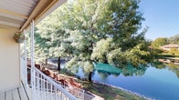 This single-story space offers a standout outdoor feature in the form of a covered balcony that delivers exceptional waterfront views. The balcony is framed by clean white railings and partially sheltered by an overhanging roofline, providing a comfortable vantage point in varying weather conditions. From this outdoor area, the view stretches across a calm body of water with striking blue-green hues, bordered by a natural stone shoreline and lush, mature trees whose canopies extend generously across the scene. The natural setting creates a serene and picturesque backdrop that is a defining characteristic of the space. The combination of the covered structure, open railing design, and proximity to the water makes the balcony a particularly inviting outdoor retreat. The mature trees lining the waterfront provide natural shade and a sense of established, organic landscaping that enhances the overall ambiance. The single-floor layout suggests ease of accessibility throughout, while the outdoor orientation toward the water ensures that natural scenery remains a central element of the living experience. The peaceful waterfront setting lends the space a relaxed, nature-connected atmosphere well-suited for quiet enjoyment of the surrounding environment.