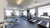Located at 100 Comet Creek Ln in Summerville, South Carolina, this fitness center is a well-equipped, single-floor facility designed to support a comprehensive workout experience. The space features a rubber-floored exercise area finished in a clean, neutral color palette with slate-blue accent walls and a light-toned ceiling fitted with bright fluorescent lighting panels and ceiling fans for optimal comfort during workouts.

The cardio section is outfitted with multiple treadmills, elliptical trainers, and a stationary bike, all positioned along the perimeter to take advantage of the large, evenly spaced windows that flood the room with natural light and offer views of the surrounding outdoors. Strength training equipment, including a weight bench and resistance machines, is arranged throughout the space to allow for efficient use of the floor area.

Two wall-mounted flat-screen televisions are positioned at strategic viewing angles, ensuring visibility from various points in the room. The layout is thoughtfully organized to provide ample space between equipment, allowing for safe and comfortable movement throughout the facility. Overall, the fitness center presents a bright, functional, and welcoming environment well-suited for daily exercise routines.