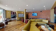 This vibrant clubroom in College Station, TX, is designed with Texas A&M spirit throughout, showcasing bold maroon-and-gold Aggie-themed murals and artwork across its walls. The main lounge area is anchored by a striking geometric tile fireplace column with a mounted flat-screen TV, surrounded by a mix of mustard yellow sectional sofas, deep red leather chairs, and colorful patterned accent seating, all set atop richly variegated hardwood floors. Exposed brick walls add an industrial warmth to the space, while large floor-to-ceiling windows along one side flood the interior with natural light and offer views of the surrounding streetscape. A shuffleboard table and arcade machines provide entertainment options, complementing the relaxed social atmosphere. A separate bar-height seating area with stools offers an additional gathering spot. Adjacent to the main lounge, a dedicated computer lab features a long built-in wood desk, multiple desktop workstations, ergonomic rolling chairs, pendant lighting, and a shared printer, set against cool blue-gray walls with patterned carpet flooring. Recessed lighting throughout and high ceilings in select areas contribute to an open, welcoming ambiance suited for both recreation and productivity.