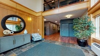 Located at 627 NE Halsey St in Portland, Oregon, this lobby and common area showcases a thoughtfully designed entrance that balances warmth and functionality. The most striking architectural feature is the soaring two-story glass wall at the entry, framed in warm wood trim and flooding the space with natural light while offering views of the surrounding greenery. Slate tile flooring in the entry transitions seamlessly into rich hardwood floors that extend throughout the interior corridors.

The main lobby area features wood-paneled wainscoting that adds a refined, welcoming character to the space. Seating is provided by a patterned accent chair paired with a sleek grey console table, accented by a decorative round mirror and wall sconce lighting. Lush potted trees add organic texture and life throughout the entry zone.

Moving deeper into the building, well-lit corridors with grey and teal accent walls guide visitors toward elevator access, marked by distinctive teal-painted doors. Framed artwork lines the hallways, contributing to a curated, community-oriented atmosphere. An upper-level mezzanine with wrought-iron railings overlooks the lobby below, reinforcing the vertical scale of the space. Overall, the design presents a polished, contemporary aesthetic rooted in natural materials and practical layout.
