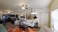 This single-floor residence offers a thoughtfully designed open-concept layout that promotes a natural flow between living spaces. The living room serves as the heart of the home, featuring neutral-toned walls and smooth ceilings accented by a ceiling fan with integrated lighting, providing both comfort and style. An elegant arched floor lamp and a bold abstract canvas painting add artistic character to the room, while a vibrant patterned area rug introduces a lively pop of color against the neutral flooring. The space is furnished with a classic white sofa, plush accent chairs, and coordinating side tables, creating a welcoming and functional seating arrangement. A graceful curved architectural detail frames the transition between the living area and the adjacent kitchen, which is visible in the background and features dark cabinetry for a striking contrast. The entry door is positioned conveniently off the living area, contributing to the efficient and accessible floor plan. Overall, the space presents a balanced combination of contemporary design elements and warm, inviting touches, making it a comfortable and visually appealing environment suited for both relaxing and entertaining.