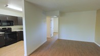 This interior space showcases a modern, minimalist design with an emphasis on openness and flow. The room features smooth, off-white walls that create a clean, bright atmosphere. The flooring is a standout element, boasting warm-toned hardwood planks that run throughout the visible area, adding a touch of natural elegance to the space.

The layout appears to be open concept, with no visible partitions in the main area. This design choice promotes a sense of spaciousness and allows for versatile use of the space. A notable architectural feature is the doorway visible in the center of the image, which leads to an adjoining room, hinting at a well-connected floor plan.

The ceiling appears to be standard height with a smooth, white finish that complements the walls. While no windows are visible in this particular view, the space is well-lit, suggesting potential natural light sources in other areas of the home.

Overall, this interior presents a blank canvas, ready for personalization. Its neutral color palette and open design make it adaptable to various décor styles and functional needs, whether for residential or commercial use.