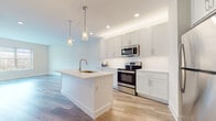 This single-floor residence showcases a clean, contemporary design with a bright and airy aesthetic carried consistently throughout. The open-concept layout connects the kitchen and living area seamlessly, creating a spacious, functional environment ideal for both everyday living and entertaining. The kitchen is a standout feature, equipped with white shaker-style cabinetry, a white subway tile backsplash, and a full suite of stainless steel appliances including a range, over-the-range microwave, and refrigerator. A central island with an undermount sink and pendant lighting above adds both practicality and visual interest to the space. Light wood-look flooring runs throughout the unit, contributing to a cohesive and modern feel. Recessed lighting and a wall-mounted mini-split system ensure comfort and well-lit interiors. The bedrooms offer neutral tones, large windows with outdoor views of natural surroundings, and ample natural light. The bathrooms are finished with white shaker vanities, white quartz countertops, LED-backlit mirrors, and wood-look flooring. One bathroom features a tub-shower combination with marble-style surround tile, while another offers a single vanity layout. The overall ambiance is fresh, clean, and move-in ready.