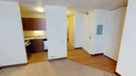 This single-floor unit features an efficient, open layout that connects the main living area to the kitchen and hallway. The living space is finished with wall-to-wall carpet in a neutral beige tone, providing a comfortable and cohesive feel. A smooth transition leads into the kitchen and entry corridor, where hardwood-style flooring adds warmth and contrast.

The kitchen is positioned to the left and is equipped with rich dark wood cabinetry, a granite-style countertop, a stainless steel sink, and a dishwasher. Under-cabinet lighting illuminates the workspace, enhancing both functionality and ambiance.

A structural wall divides the kitchen from the entry corridor, which features the unit's entry door alongside an electrical panel box. A hallway visible beyond the dividing wall leads further into the unit, suggesting access to additional rooms such as a bedroom or bathroom. Flush-mount ceiling lighting provides consistent illumination throughout the space. The overall design is practical and straightforward, with a clean, well-maintained interior finish.