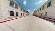 This exterior front view showcases a well-appointed multi-unit complex consisting of two parallel, two-story buildings that face each other across a broad, open concrete drive. The buildings feature a refined classical architectural style, clad in light-toned stucco and stone-block detailing that lends a clean, professional appearance. Each structure is lined with individually numbered, white panel entry doors — each flanked by wall-mounted lantern-style sconces — providing clearly defined, private access points for each unit. Large double-hung and multi-pane windows, trimmed with decorative moldings, run along both levels, offering abundant natural light. Arched entryways with columned detailing punctuate the ends of both buildings, adding a distinguished architectural accent. The central courtyard-style drive is finished in smooth concrete and lined with bold red-painted curbs designating fire lanes. Minimal landscaping with gravel beds runs along the base of each building, maintaining a tidy, low-maintenance appearance. The open-sky setting allows for expansive views, and the overall layout promotes ease of access and clear circulation throughout the property. The complex presents a polished, orderly exterior well-suited for professional or multi-tenant use.