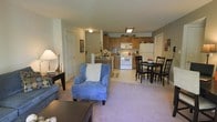 This single-floor residence showcases an open-concept layout that seamlessly connects the living room, dining area, and kitchen into one cohesive, functional space. The living room is carpeted and comfortably furnished, featuring a plush blue armchair with a decorative pillow, a sofa, and a coffee table, creating a relaxed and inviting atmosphere. Soft, warm lighting from a table lamp complements the neutral wall tones throughout.

Adjacent to the living area, a dining space accommodates a dark-finished table with matching chairs, suitable for everyday meals or casual gatherings. A bowl of fruit on the table adds a homey touch.

The kitchen is positioned at the rear of the open floor plan and features warm wood-toned cabinetry, a white electric range, a white refrigerator, and overhead lighting that illuminates the workspace effectively. The kitchen floor transitions from carpet to a light-toned hard surface, clearly delineating the cooking and prep area from the living spaces. A small decorative rug marks the transition zone. Two doors visible along the left wall suggest access to additional rooms or storage, adding practical functionality to the overall layout.