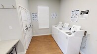 This single-story shared amenity facility is designed to serve a residential community, offering dedicated laundry and secure personal storage areas within a clean, well-maintained environment. The laundry room is equipped with a row of top-loading washing machines and stacked dryer units, providing ample capacity for multiple users. Instructional signage for machine operation is posted throughout, ensuring ease of use. A window with blinds allows natural light into the laundry area, contributing to a bright and functional atmosphere. Wood-look flooring runs throughout the space, adding warmth to the otherwise utilitarian setting, while neutral gray walls keep the environment clean and understated.

An adjoining area serves as a secure storage room, lined with a substantial bank of gray metal lockers arranged in double-tier rows. This room is accessible via glass-panel double doors that open to the outdoors, providing convenient entry. A separate entry area with additional exterior access features a folding table, wall-mounted hooks, and a window, functioning as a transitional or utility space. The facility is thoughtfully organized to support day-to-day community needs efficiently.