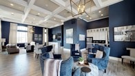 This single-floor space makes a striking first impression with its soaring coffered ceilings detailed in crisp white millwork, lending architectural depth and a sense of grandeur to the open layout. Bold navy blue walls create a dramatic yet sophisticated backdrop that anchors the interior design and ties together the various functional areas seamlessly.

The main lounge area is furnished with plush navy velvet wingback chairs accented by nailhead trim, arranged around a central coffee table to encourage conversation. A geometric brass and glass chandelier serves as a focal point overhead, complementing the warm-toned hardwood-style flooring that runs throughout.

The space flows naturally into a dining area, where neutral upholstered chairs and a coordinating chandelier maintain the refined aesthetic. Adjacent to these living areas, a well-appointed kitchen features tall white cabinetry with glass-front upper cabinets, a stainless steel refrigerator, and a raised bar-height counter with seating, offering both functionality and style.

Contemporary artwork adorns the walls, and layered lighting throughout enhances the inviting, upscale ambiance of this cohesive, open-concept space.