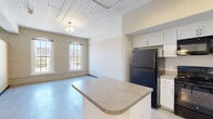 Located at 204 Wayne Ave in Dayton, Ohio's vibrant Wayne Avenue corridor, this single-floor unit showcases an impressive blend of historic industrial character and contemporary updates. Exposed painted brick walls, polished concrete floors, and tongue-and-groove wood ceilings create a distinctive loft aesthetic throughout the space.

The open-concept layout connects the living area and kitchen seamlessly. The kitchen is well-appointed with white shaker-style cabinetry, neutral countertops, black appliances including a refrigerator, electric range, built-in microwave, and dishwasher, as well as a central island that provides additional prep and seating space. Track lighting and pendant fixtures illuminate the area effectively.

The living space is generously sized and filled with natural light, courtesy of tall arched double-hung windows that frame views of the surrounding Dayton streetscape. Ceiling fans add comfort throughout. A dedicated bedroom features its own arched window, painted brick accent walls, and a ceiling fan, offering a bright and airy retreat.

The bathroom is clean and functional, featuring a walk-in shower, a white vanity with a gray countertop, and polished concrete flooring consistent with the rest of the unit. The overall ambiance is modern-industrial, with thoughtful finishes that honor the building's architectural heritage.