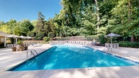 Located at 4401 Hamptonridge Drive in Charlotte, NC, this two-floor property is centered around a generously sized outdoor swimming pool that serves as the focal point of the community. The pool itself is notably spacious, with clearly marked depth indicators and lane dividers, making it suitable for both leisure and fitness swimming. Surrounding the pool is a wide, well-maintained concrete sundeck lined with rows of lounge chairs and patio umbrellas, providing ample space for relaxation and sunbathing. A white pergola structure anchors one end of the deck, offering a shaded gathering area complemented by outdoor furniture. Adjacent to the pool, a charming cabana-style building with a pitched roof, chimney, and warm siding houses what appears to be restroom and support facilities, adding both function and architectural character to the area. The entire pool area is embraced by mature, towering trees and manicured hedgerows, creating a private, resort-like atmosphere that feels naturally secluded. The lush greenery provides a verdant backdrop that enhances the tranquil ambiance of the outdoor space. Whether relaxing poolside or enjoying the shade of the pergola, this property offers a well-appointed outdoor retreat in the heart of Charlotte.
