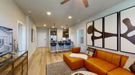 This thoughtfully designed single-floor residence showcases a seamless open-concept layout connecting the kitchen, dining, and living areas. The kitchen is equipped with stainless steel appliances, dark wood cabinetry, a subway tile backsplash, and a granite-topped island with seating for multiple bar stools — ideal for casual dining and entertaining. Pendant lighting overhead adds a modern touch, while light hardwood flooring flows throughout the main living areas, creating a warm and cohesive aesthetic.

The living room is generously proportioned, featuring a ceiling fan, recessed lighting, and large windows and a glass door that allow natural light to fill the space. The room easily accommodates substantial seating and is accented by bold, large-format wall art.

The bedroom offers a comfortable, carpeted retreat with ample natural light from well-sized windows dressed in layered blinds and curtains. A ceiling fan provides additional comfort.

The bathroom is finished with dark granite countertops, dark wood vanity cabinetry, light hardwood-style flooring, and a tiled tub-shower combination. A second bathroom provides similar finishes in a clean, functional layout. Throughout, the neutral color palette and modern design elements contribute to a cohesive, contemporary living environment.