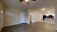 This single-floor space presents a well-organized and functional layout with a clean, move-in-ready aesthetic. The main living area is finished with rich, dark hardwood-style flooring that extends throughout, providing both durability and visual warmth. Neutral beige walls create a versatile backdrop suitable for a variety of interior styles, while white baseboards offer a crisp contrast that enhances the overall finish of the space.

A ceiling fan with wood-toned blades is centrally mounted, contributing to both air circulation and a subtle decorative touch. The room features two doors along one wall — one leading to a walk-in closet, which is visible through the open doorway and appears to offer generous storage with built-in shelving or rod space. The second door provides access to an additional room or utility area.

Toward the rear of the space, a transition to lighter flooring is visible, suggesting a distinct adjoining area, potentially a kitchen or secondary room. Recessed and flush-mounted lighting fixtures provide consistent illumination throughout. The overall ambiance is bright, tidy, and practical, reflecting a well-maintained and thoughtfully designed interior.