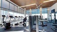 This contemporary fitness center is defined by its striking floor-to-ceiling glass walls, which flood the space with abundant natural light and offer open views of the surrounding cityscape. The warm wood-paneled ceiling adds a refined architectural contrast to the sleek, modern equipment below, creating a polished yet inviting atmosphere. Large industrial-style ceiling fans are mounted overhead, ensuring consistent airflow throughout the space during workouts. The layout is thoughtfully organized, with a row of treadmills positioned along the windowed perimeter, allowing users to enjoy outdoor views while exercising. Strength and resistance machines are arranged in the central floor area, providing a well-rounded selection of workout options. White cylindrical columns punctuate the space at intervals, contributing to its clean aesthetic without disrupting the open, expansive feel of the floor plan. Durable rubber flooring covers the entire workout area, offering both safety and practicality. The combination of natural light, high-quality equipment, and considered design details makes this fitness center a functional and visually appealing space suited for a variety of exercise routines.