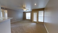 This single-floor residence offers a clean, functional layout designed for comfortable everyday living. The main living area is a generously sized room finished with wall-to-wall carpeting in a neutral tan tone, providing a warm and inviting atmosphere. Smooth, light-colored ceilings are fitted with a ceiling fan and integrated light fixture, ensuring year-round comfort and overhead illumination. A recessed can light adds a modern touch and enhances brightness throughout the space. The room benefits from abundant natural light, courtesy of a large window and a glass-paned exterior door with an adjacent sidelight panel, both dressed with horizontal blinds for privacy and light control. The door provides direct outdoor access, making the space feel connected to the exterior. Neutral taupe-toned walls complement the carpeted floors and white trim, creating a cohesive and understated aesthetic throughout. The clean lines and open layout of the living area make it highly versatile, easily accommodating a variety of furniture arrangements. The overall ambiance is bright, open, and move-in ready, offering a welcoming environment suited to a range of lifestyles.