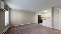 This single-floor unit offers a practical and well-organized layout with a seamless flow between living and functional spaces. The open living area is finished with wall-to-wall carpet in a neutral tone, providing a warm and comfortable foundation throughout. Neutral-toned walls and white baseboards contribute to a clean, understated aesthetic.

The kitchen is positioned adjacent to the living area and features white shaker-style cabinetry with brushed nickel hardware, marble-patterned laminate countertops, and a stainless steel sink. Black appliances — including a refrigerator, electric range with overhead exhaust hood, and dishwasher — offer a bold contrast to the light cabinetry. The flooring transitions to a light tile, complementing the bright, functional workspace.

A dedicated bedroom area includes two windows fitted with horizontal blinds, a wall-mounted air conditioning unit, and baseboard heating — offering year-round climate control. Natural light enters through multiple windows, brightening the space throughout the day.

The bathroom is finished with tile flooring, a white vanity with a mirror cabinet, and clean neutral walls. A dedicated closet area houses a stacked washer and dryer unit alongside built-in shelving, adding convenient in-unit laundry storage.