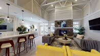 Located at 2000 Crossroads Manor Ct in Cary, NC, this single-floor community clubhouse is a well-appointed gathering space designed to serve a variety of social and professional needs. The centerpiece of the main lounge is a dramatic vaulted ceiling with exposed white truss detailing, creating an airy, expansive atmosphere. A floor-to-ceiling stacked stone fireplace anchors the seating area, which is furnished with plush sofas in bold yellow and gray tones, complemented by light wood-look tile flooring throughout.

White structural columns define distinct zones within the open-plan layout, guiding visitors from the welcoming entrance — with its large glass French doors and tropical greenery — into the main gathering area. A bar-height counter with seating provides a casual lounge spot beneath pendant lighting and a decorative mirror.

A quieter lounge wing features soft teal-painted walls with classic wainscoting, an arched window, and a mix of seating arrangements including a navy velvet sofa and light blue wingback chairs, making it ideal for relaxed conversation or individual work. A wall-mounted television and workstation desk add functional versatility to this area.

A dedicated conference room offers a rustic wood table, leather chairs, a whiteboard, and bright windows with outdoor views. Additional amenities include a dual-level water fountain and bottle-filling station. The space is thoughtfully designed to balance comfort, style, and community functionality.
