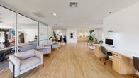 Located at 6336 S 82nd E Ave in Tulsa, Oklahoma, this well-appointed single-floor commercial space offers a thoughtfully designed, open-plan interior finished with light wood flooring throughout. The bright, white-painted walls and recessed lighting create a clean, contemporary aesthetic that feels both professional and welcoming.

The main open area serves as the central hub of the space, accommodating multiple workstations, including a dedicated desk area equipped with computer monitors, as well as a private office nook featuring a mid-century style wood desk and guest seating. A comfortable lounge area with a sofa, armchairs, and a round coffee table provides an inviting space for meetings or informal gatherings, accented by lush indoor plants and modern wall art.

A glass-partitioned section at one end of the space houses fitness equipment, offering a clearly defined area without disrupting the open flow of the interior. Toward the back, a functional kitchenette with light wood cabinetry provides practical amenity support. Large glass entry doors along the front facade flood the space with abundant natural light while offering views of the well-maintained outdoor greenery, further enhancing the bright and airy ambiance throughout.