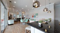 This single-story clubhouse offers a welcoming and well-appointed communal space designed for both relaxation and socializing. The open-concept layout seamlessly connects a dining area, lounge zone, and a kitchen counter space, creating a cohesive and functional environment.

The dining area is furnished with round tables and distinctive mint green lattice-back chairs, adding a playful, coastal charm to the space. Overhead, polished chrome dome pendant lights serve as eye-catching focal points, complementing the recessed lighting that illuminates the room evenly. Crown molding runs along the perimeter of the white ceiling, lending a refined architectural touch.

The kitchen counter features a bold dark granite surface paired with white cabinetry and white bar stools, offering casual seating and a practical workspace. Potted greenery on the counter adds a fresh, natural accent.

A lounge area in the background provides additional seating with comfortable sofas, while colorful framed artwork — featuring figures in vibrant dresses — decorates the walls, enhancing the cheerful, resort-inspired aesthetic. Warm wood-look flooring flows throughout, tying the entire space together with a clean, modern finish.