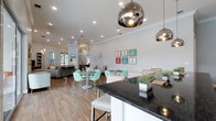 This single-story clubhouse offers a welcoming and well-appointed communal space designed for both relaxation and socializing. The open-concept layout seamlessly connects a dining area, lounge zone, and a kitchen counter space, creating a cohesive and functional environment.

The dining area is furnished with round tables and distinctive mint green lattice-back chairs, adding a playful, coastal charm to the space. Overhead, polished chrome dome pendant lights serve as eye-catching focal points, complementing the recessed lighting that illuminates the room evenly. Crown molding runs along the perimeter of the white ceiling, lending a refined architectural touch.

The kitchen counter features a bold dark granite surface paired with white cabinetry and white bar stools, offering casual seating and a practical workspace. Potted greenery on the counter adds a fresh, natural accent.

A lounge area in the background provides additional seating with comfortable sofas, while colorful framed artwork — featuring figures in vibrant dresses — decorates the walls, enhancing the cheerful, resort-inspired aesthetic. Warm wood-look flooring flows throughout, tying the entire space together with a clean, modern finish.