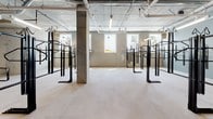 This single-floor bike storage facility is designed with functionality and capacity in mind, offering a well-organized environment for bicycle parking and security. The space is divided into distinct zones, each serving a specific storage purpose.

The primary storage area features rows of freestanding Saris bike racks arranged in a symmetrical layout, maximizing the number of bicycles that can be accommodated. Natural light filters in through ground-level windows, providing visibility throughout the space. A secondary area along the perimeter walls is lined with wall-mounted Saris bike racks, offering an efficient vertical storage solution that conserves floor space.

The industrial aesthetic is consistent throughout, with exposed concrete ceilings revealing an extensive network of mechanical pipes and ductwork in contrasting colors. Polished concrete floors provide a durable, low-maintenance surface suited to high-traffic use. Concrete columns punctuate the open floor plan, adding structural character while supporting the ceiling grid above.

A seating area with a built-in concrete bench and modern chairs provides a functional rest space, accompanied by a decorative mural on the adjacent wall. A chain-link enclosed utility area houses mechanical systems. Access is provided via a secured door leading to additional building amenities.