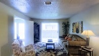 This single-story space centers around a comfortable living room that serves as the heart of the home. The room features a textured popcorn ceiling with a centrally located HVAC vent, contributing to a classic residential feel. Natural light pours in through a large sliding glass door dressed with vertical blinds, creating a bright and airy atmosphere while offering potential outdoor access. The living room is generously sized, accommodating a plush sectional sofa accented with decorative throw pillows, a carved wooden coffee table, and a patterned area rug that anchors the seating arrangement. Decorative elements include tall bamboo-style plants in the corners, a dark wood bookcase along one wall, and wall art that adds personality to the space. An additional doorway on the left side of the room provides connectivity to other areas of the home. The neutral wall tones complement the warm, earthy furnishings, creating an inviting and functional living environment suitable for everyday use and casual entertaining.