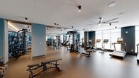 Located at 2000 Schindler Drive in South Amboy, NJ, this fitness center is a well-appointed exercise facility designed to support a full spectrum of workout routines. The space is housed on a single floor and offers an expansive, open layout finished with durable rubber flooring throughout, providing both comfort and functionality.

The facility is divided into distinct workout zones. A dedicated cardio area lines the perimeter with floor-to-ceiling windows, filling the space with abundant natural light and an open, airy ambiance. This section is equipped with multiple treadmills, stationary bikes, and elliptical machines, each featuring integrated display screens. Large ceiling fans complement the ventilation system overhead.

The strength training area is equally comprehensive, featuring a multi-station cable machine, a power rack with an integrated lifting platform, barbells, and an extensive dumbbell rack. Mirrored walls amplify the sense of space and allow for proper form monitoring. A dedicated free-motion and functional training section includes stair climbers and additional cable equipment.

A separate studio area offers a smooth, open floor ideal for stretching, yoga, or group exercise, complete with a large wall-mounted display screen and full-length mirrors. The overall design palette of deep blue-gray accent walls, white ceilings, and warm timber details creates a modern, motivating environment throughout.